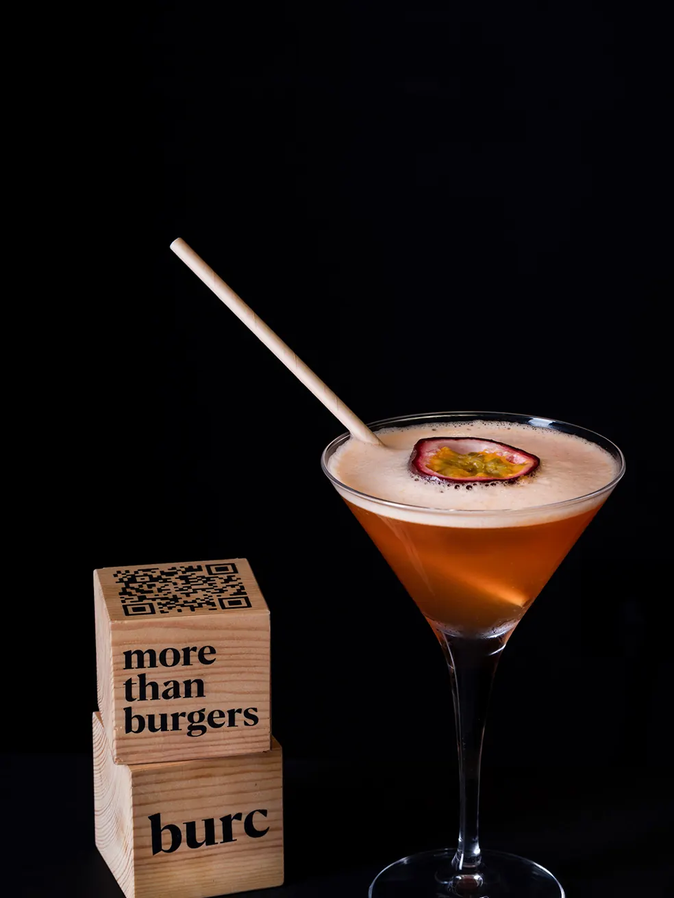 Pornstar Martini en copa, junto a un cubo de madera con el logotipo burc