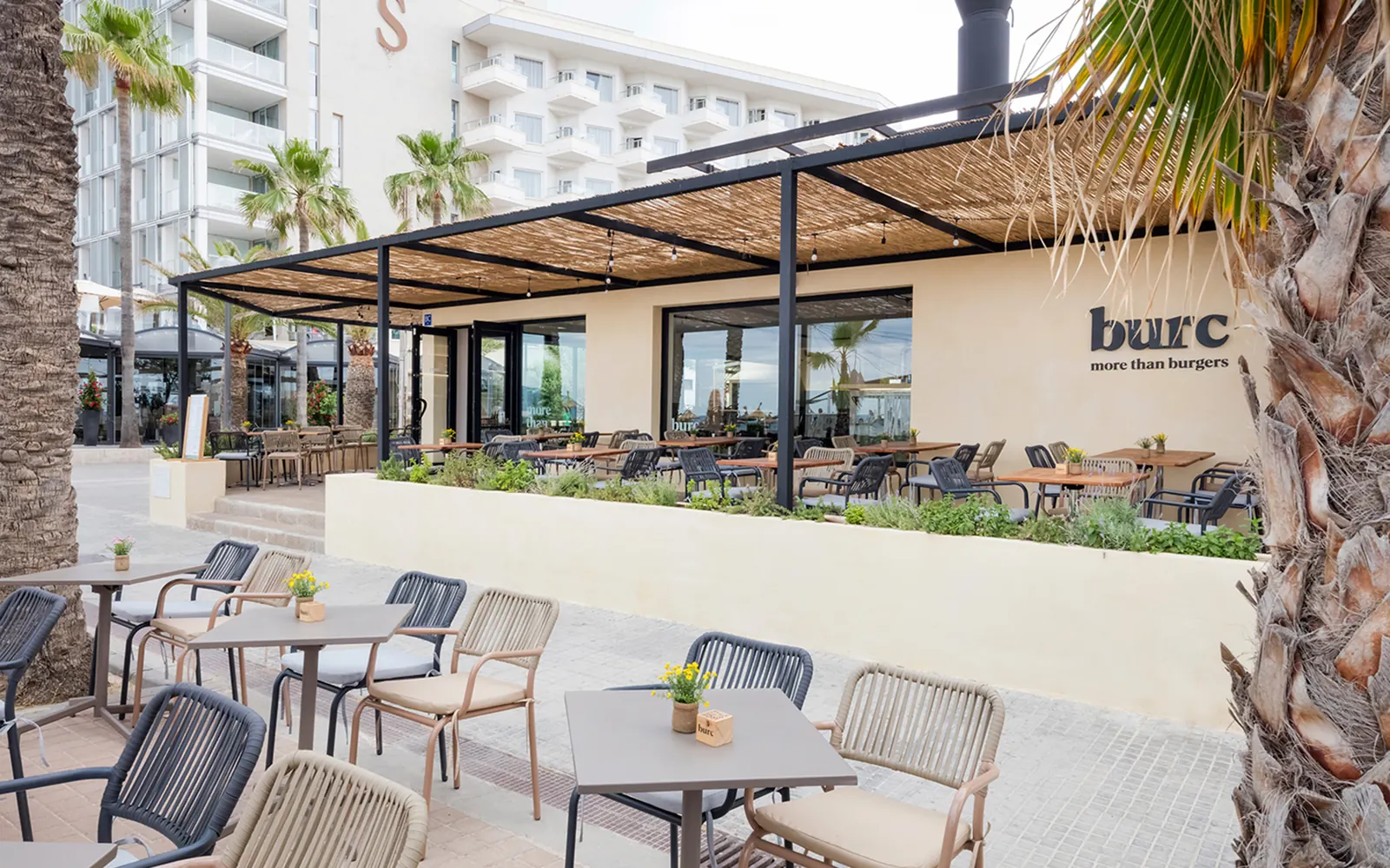 Terraza exterior con mesas y sillas de fibra, palmeras y el hotel vecino al fondo
